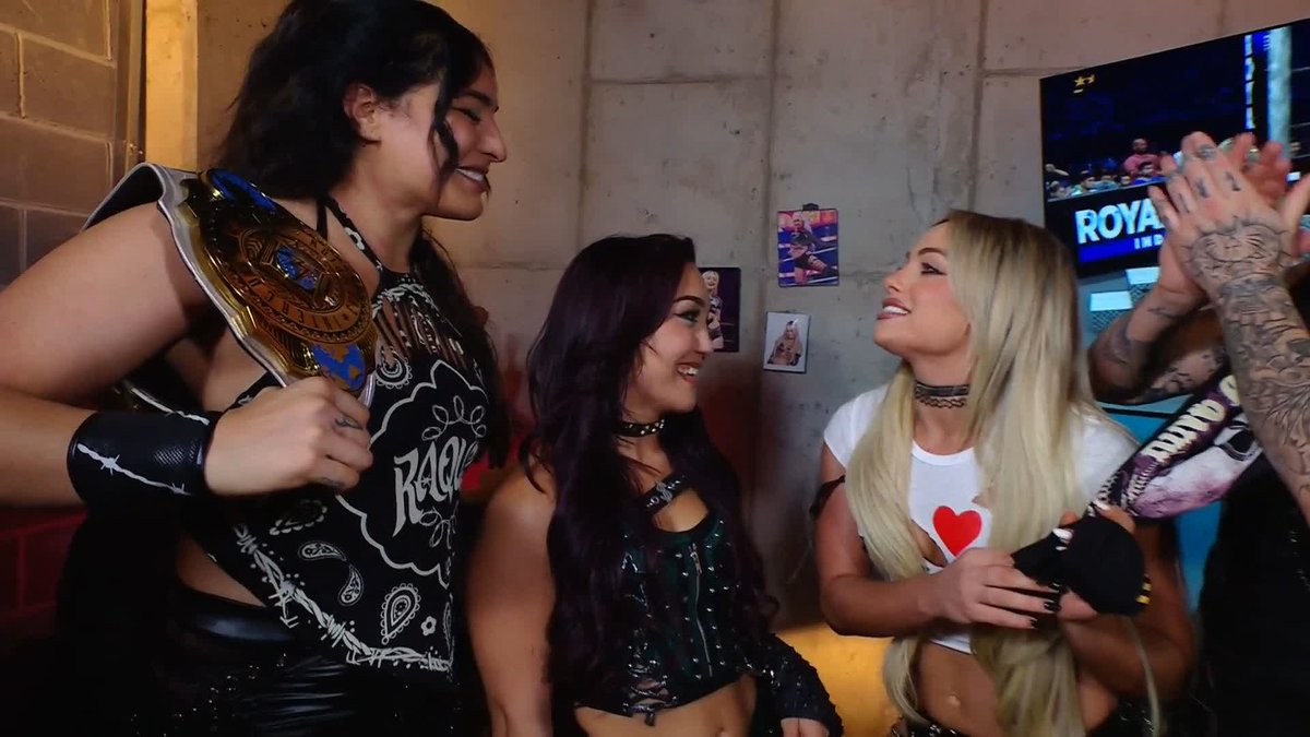 The photo captures three women smiling warmly in what appears to be a backstage wrestling setting, with one prominently holding a championship belt, while a TV screen in the upper right displays "ROYAL RUMBLE" text. The scene reflects the celebratory backstage moments that wrestlers like jd_mcdonagh and Joe Hendry share with fellow performers after major events.