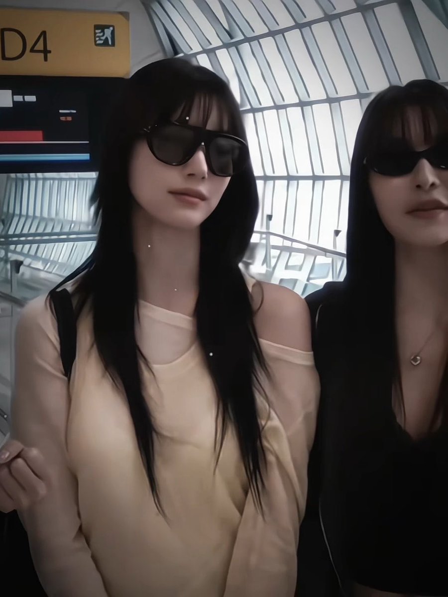 Two women wearing large black sunglasses pose at what appears to be an airport, with a yellow gate sign reading 'D4' visible in the background.