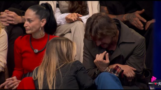 A man with long brown hair looks down while seated next to a woman in a red dress on what appears to be a televised reunion or interview set, with the hashtag "#AFazenda" visible, suggesting this is from the Brazilian reality show "A Fazenda" where contestants are sharing their thoughts or reactions.