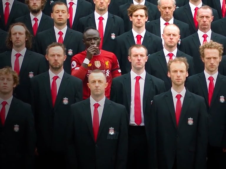 A man in a red Liverpool soccer jersey stands out among a group of men dressed in identical dark suits with red ties.