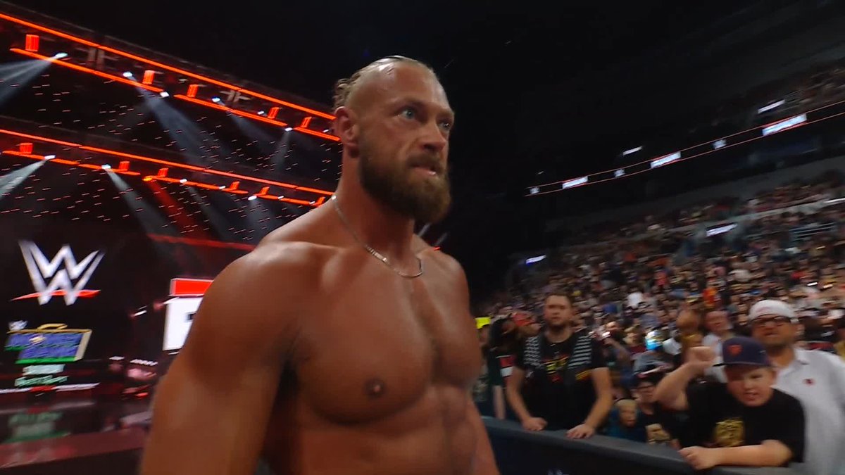 A muscular, bearded wrestler with a top knot walks through a WWE arena packed with fans, with the WWE logo and "RAW" visible on the entrance stage screen in the background. The image captures the imposing presence of Big Cass following his attack on Dragon Lee and Rey Mysterio.