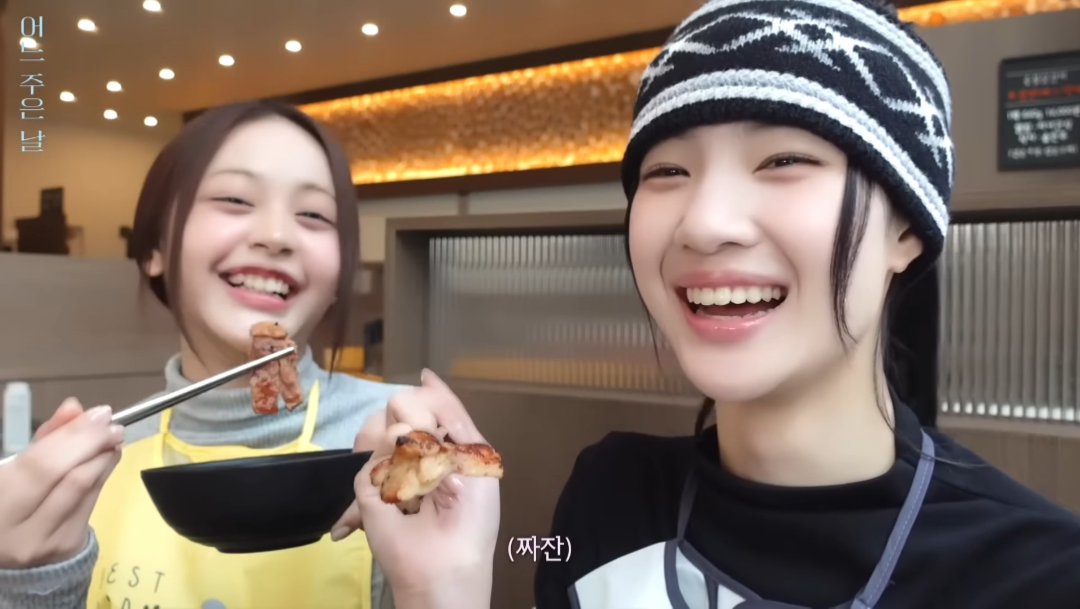 Two young women smile and laugh while eating food with chopsticks in a restaurant setting, wearing yellow aprons; Korean text including '뭐먹는지발' and '(짜잔)' is visible on screen.