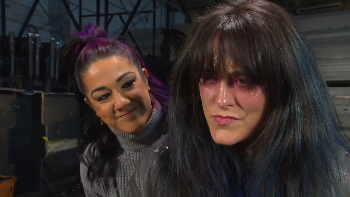 The image depicts WWE wrestler Bayley smiling subtly in the foreground, while a figure wearing a dark wig with bruised makeup around the eyes stands in the background—visually representing the post's caption that "Bayley is in the head of Lyra Valkyria" on WWE RAW. The contrast between Bayley's confident smirk and the battered, disheveled appearance of the second figure suggests a psychological rivalry.