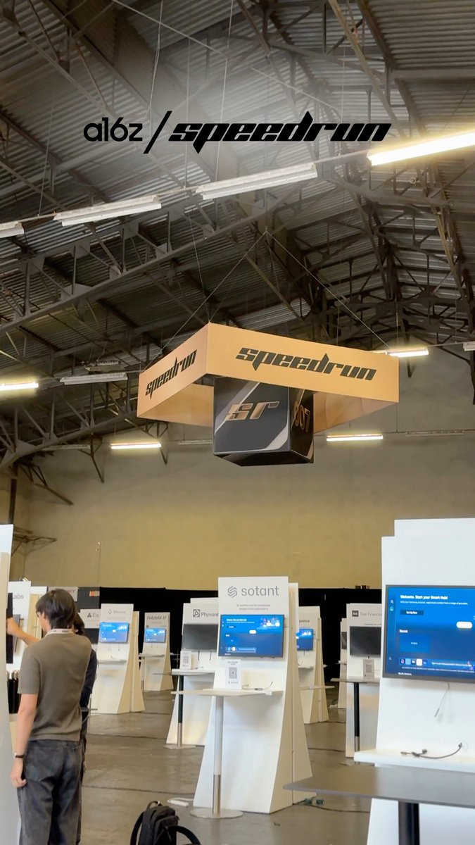 Convention or trade show venue featuring a hanging 'a16z/speedrun' sign and cardboard 'speedrun' display, with multiple tech company booths below.