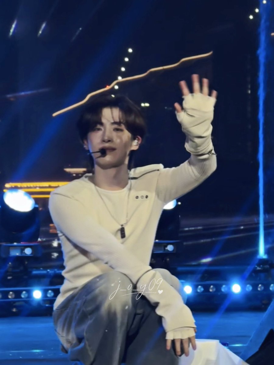 A young male performer with a headset microphone poses on stage in a cream-colored sweater and light pants, his hands wrapped in bandages, with a 'j_o_y09' watermark.