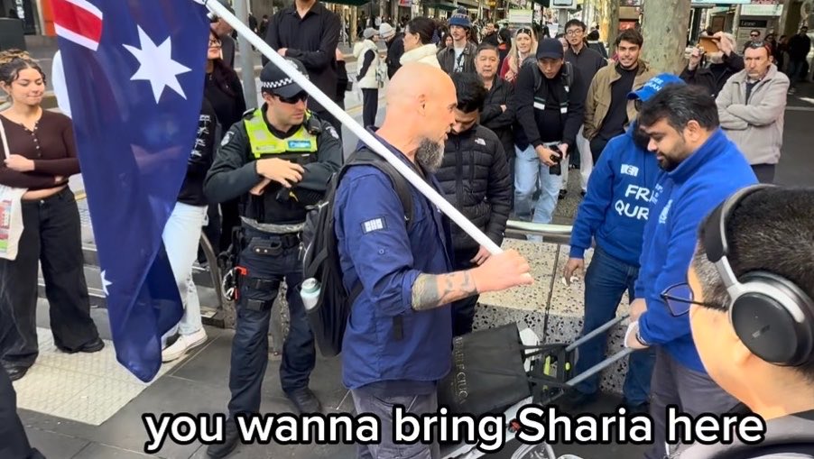 A man holds an Australian flag in a public area with a police officer and crowd nearby, while a man in a blue 'FREE QURAN' hoodie faces him; subtitle reads 'you wanna bring Sharia here'.
