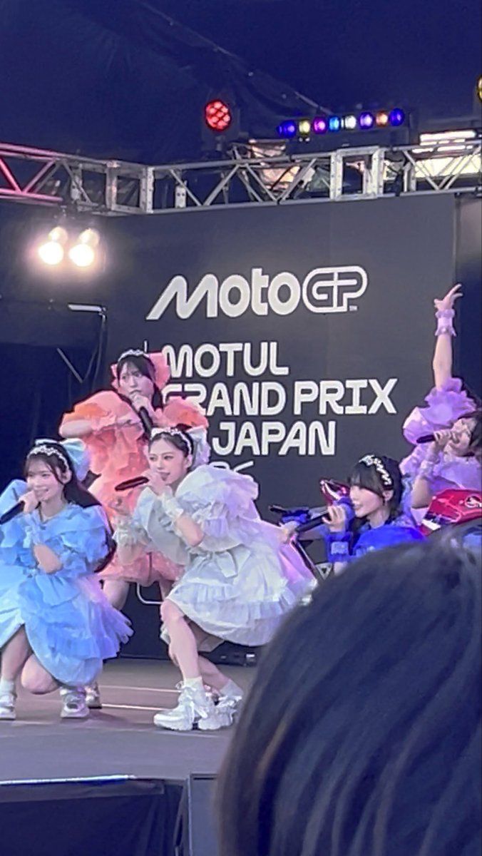 Performers in pastel blue, pink, and purple dresses singing on a stage at the 'MotoGP MOTUL GRAND PRIX JAPAN' event.