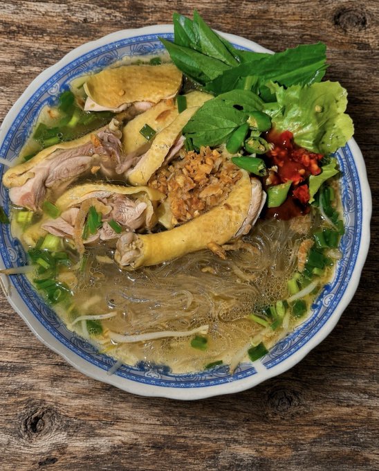 A bowl of chicken noodle soup served in a blue and white patterned bowl, garnished with fresh herbs, green onions, and red chili peppers.