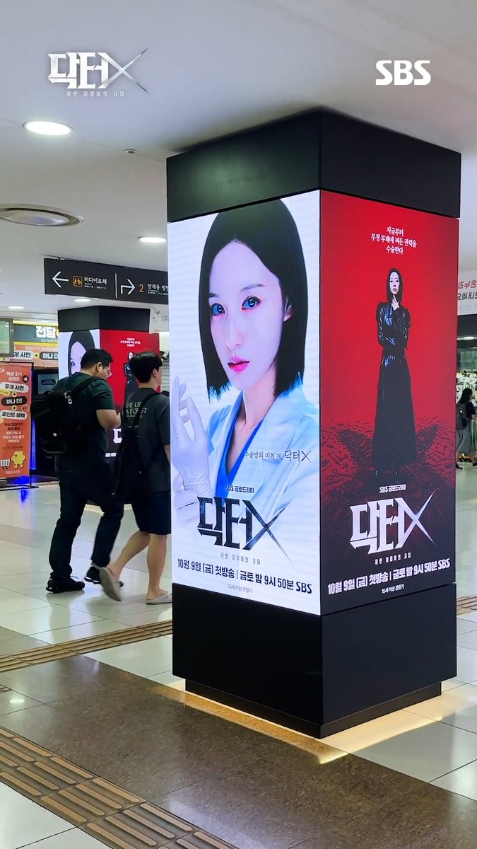 An SBS advertisement billboard in a subway station features the Korean drama 'The Escape of the Seven' (7인의 탈출), airing October 9, with a woman in a white coat on one side and a woman in a black dress on the other.