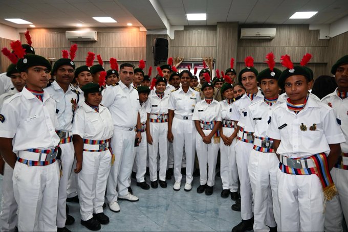 A group of NCC cadets in white uniforms with red-plumed berets stand in formation inside a room with air conditioning units.
