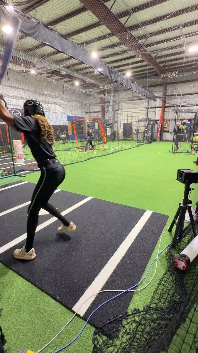 A batter in black athletic gear swings at an indoor baseball training facility with green turf and netting.