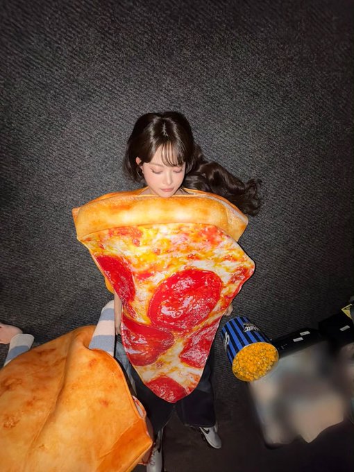 A person sitting against a gray wall hugging a large plush pillow designed to look like a slice of pepperoni pizza.