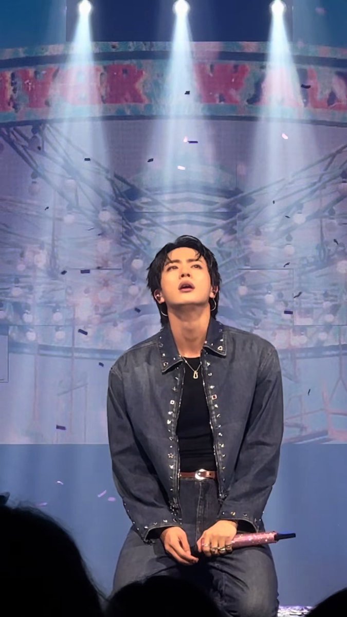 A male K-pop performer in a studded denim jacket kneels on stage holding a pink microphone, with confetti falling around him under bright spotlights.