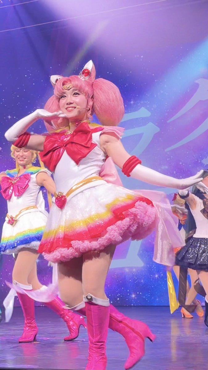 A performer dressed as Chibi Moon (Sailor Chibi Moon) from Sailor Moon in a pink and white costume with cat-ear headband, performing on stage with other Sailor Moon cosplayers in the background.