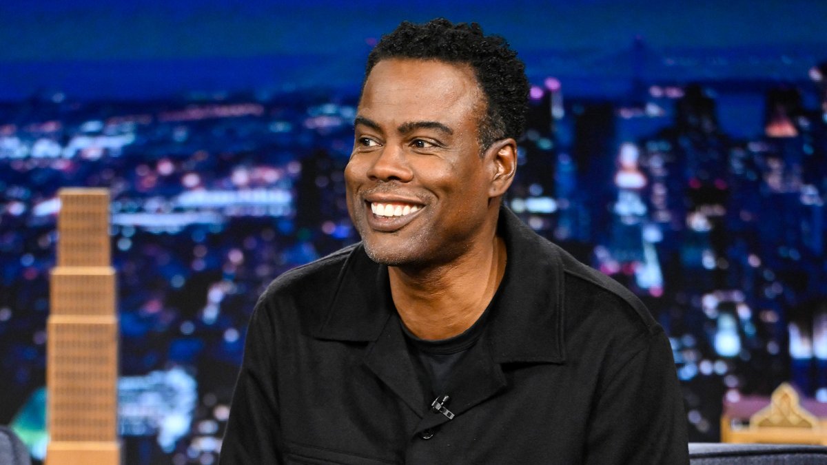 Chris Rock smiles during an appearance on The Tonight Show Starring Jimmy Fallon, appearing as a guest on the late-night program against a backdrop of a nighttime city skyline.