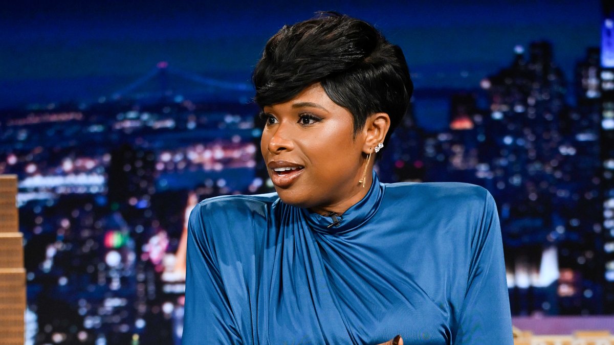 Jennifer Hudson appears as a guest on "The Tonight Show Starring Jimmy Fallon," wearing an elegant blue high-neck blouse with her hair styled in a short pixie cut, seated against the show's iconic New York City skyline backdrop.