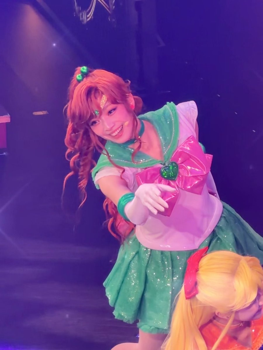 A performer on stage in a Sailor Jupiter cosplay costume (green and pink) with a glowing forehead jewel, smiling and posing toward the camera.