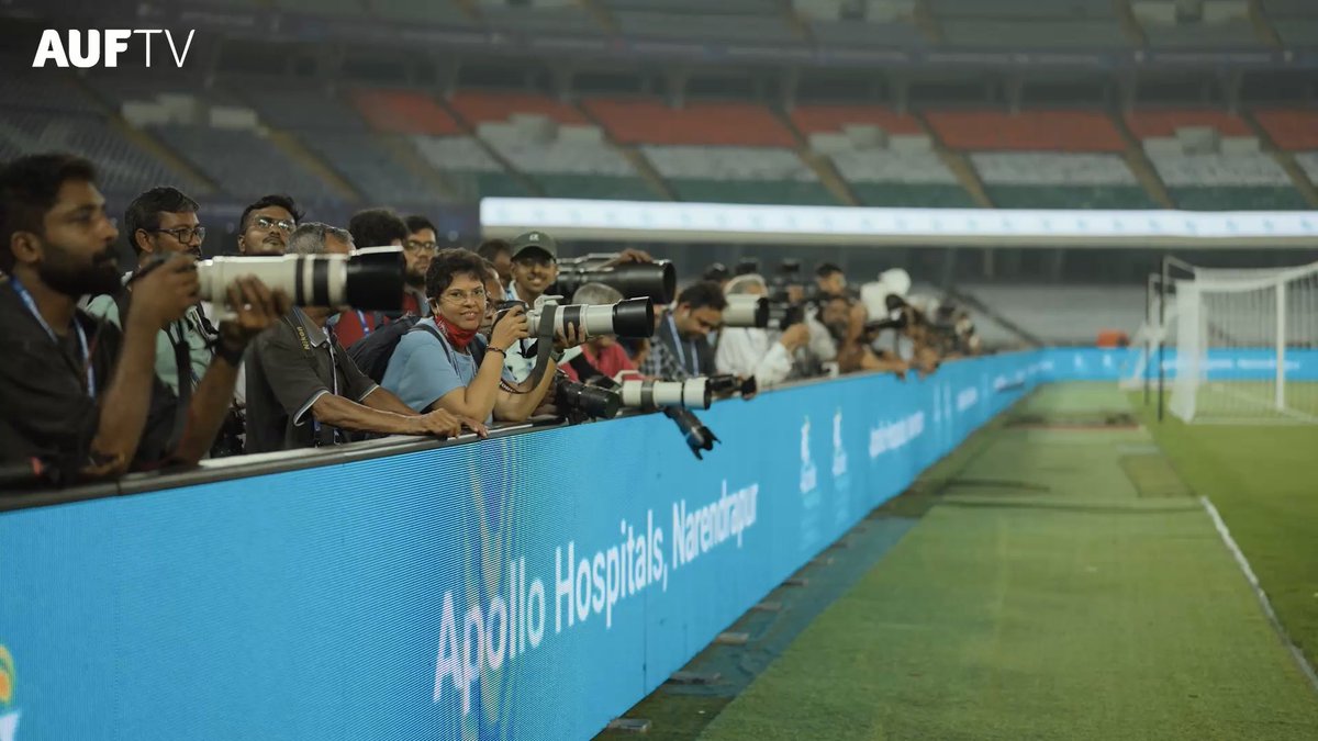 A row of photographers with long telephoto lenses line the pitchside at a stadium, leaning over an advertising hoarding that reads "Apollo Hospitals, Navasariapuram," with the "AUFTV" logo visible in the top left corner. The setup of professional-grade equipment and branded sponsorships visually counters the post's complaint about poor camera quality from the BCCI.