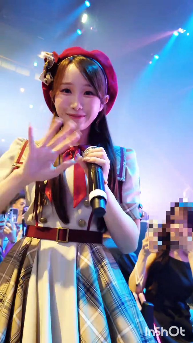 A young woman in a plaid dress and red beret performing on stage with a microphone, making a hand gesture towards the camera.
