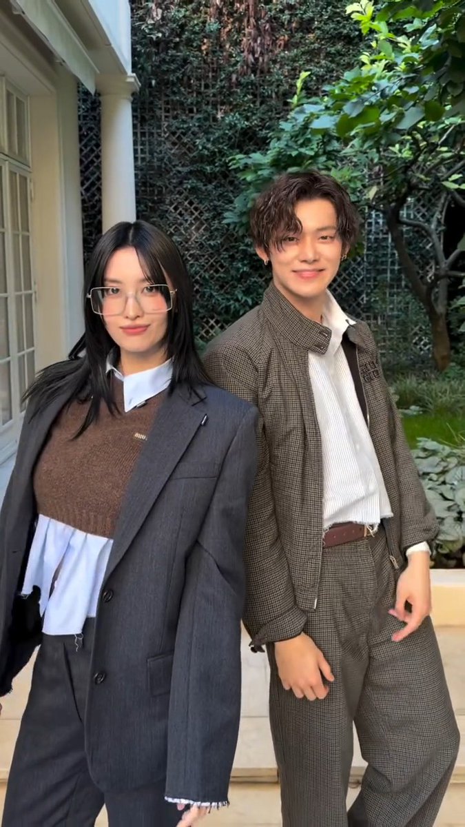 Two young people posing back-to-back outdoors, with the woman wearing glasses and a grey blazer over a brown sweater, and the man in a checkered brown suit.