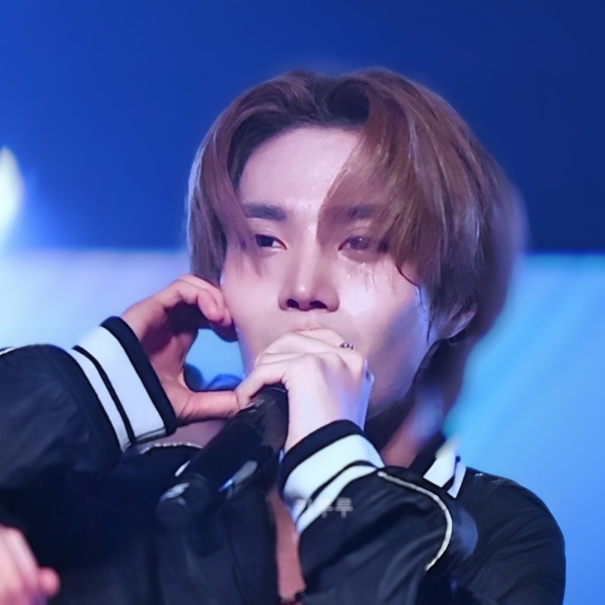 A close-up of a male K-pop performer with light brown hair, holding a microphone and making a heart gesture with his hands, wearing a black jacket with white trim.