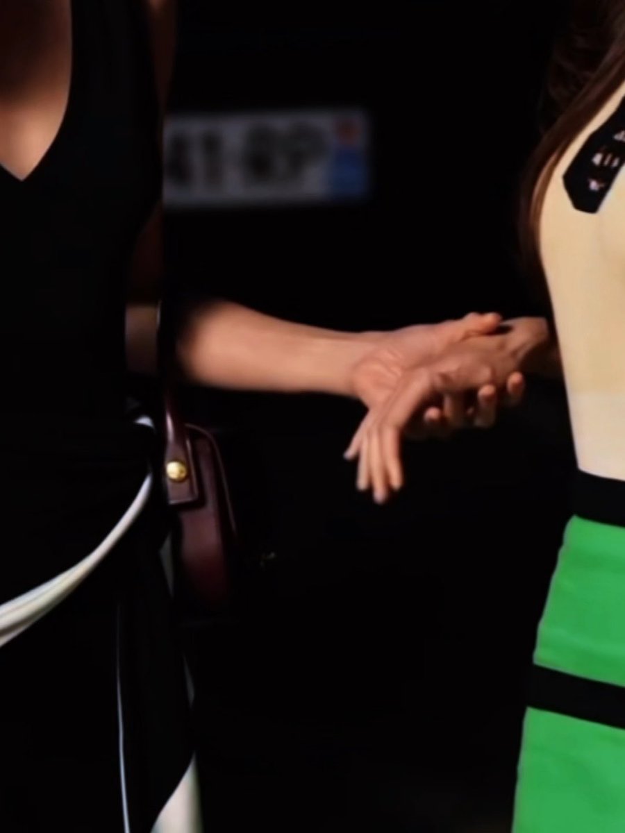 A close-up of two people whose hands are clasped together, one wearing a black outfit with a burgundy bag strap and the other wearing a white top with a green skirt.