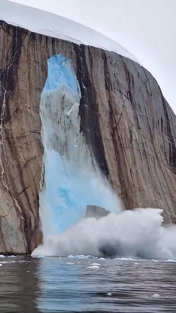 A massive chunk of ice dramatically calves off the steep, rocky face of Greenland's Petermann Glacier, crashing into the water below and sending up a huge plume of spray and mist. The towering blue-tinged ice formation dominates the frame against the dark cliff walls, illustrating the scale of the glacier's collapse.