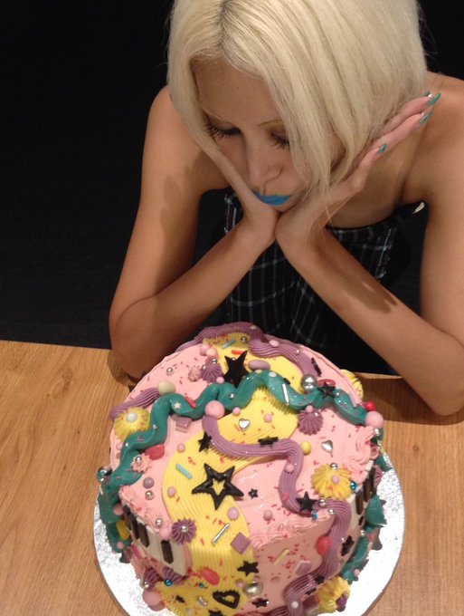 A blonde woman rests her chin on her hands while looking down at a decorated pink and yellow birthday cake adorned with stars and colorful piping.