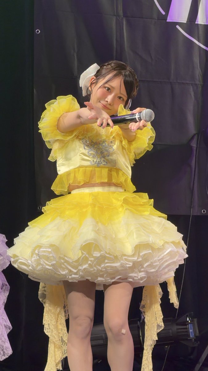 Photo of a young female performer on stage wearing a ruffled yellow and white frilly dress, extending her arms toward the camera while holding a microphone, with a white bow in her hair.