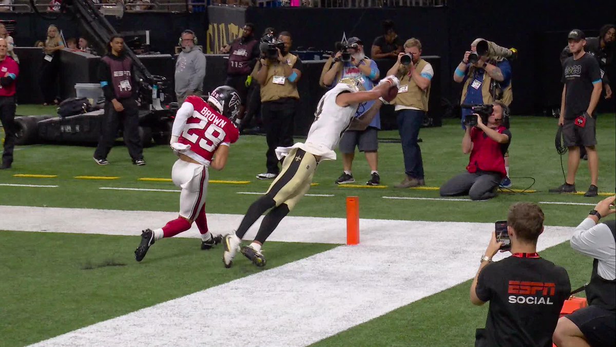 A football player in a white and black uniform makes an acrobatic catch near the pylon in the end zone, leaping over defender #29 "Brown" of the Atlanta Falcons, while numerous photographers capture the moment, including an ESPN Social staff member, bringing Devaughn Vele's spectacular grab to life.