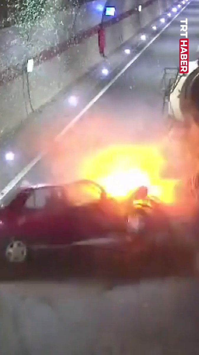 A red car is engulfed in flames on a road at night, with the TRT HABER logo visible in the upper right corner, capturing the aftermath of a fatal collision with a concrete mixer in a Trabzon tunnel where the driver lost their life.