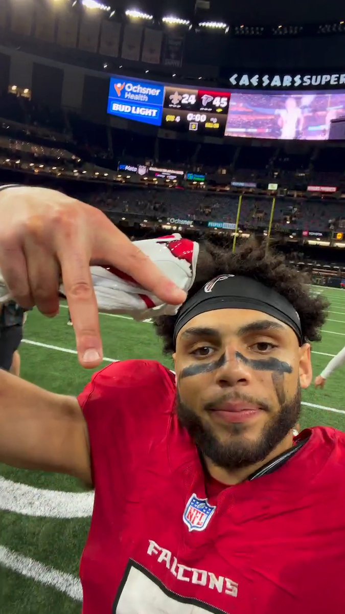 An Atlanta Falcons player wearing a red jersey with "FALCONS" and an NFL logo takes a celebratory selfie on the field at the Caesars Superdome, flashing a peace sign with black eye black smeared across his face. The scoreboard behind him shows a Saints-Falcons final score of 24-45, with the Falcons logo displayed prominently after their lopsided victory.