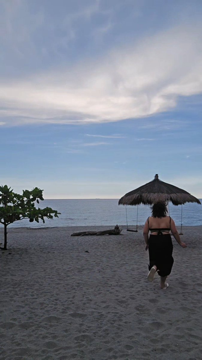 A woman in a black dress walks on a sandy beach toward the ocean at sunset, with a thatched-roof swing hut and a small tree visible.