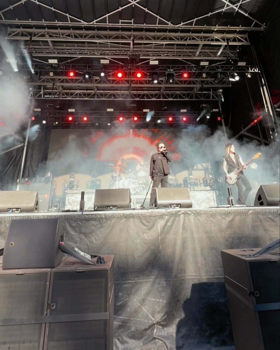 A concert stage with dramatic red lighting and truss rigging overhead, showing two performers — a vocalist with a microphone and a bassist — performing amid theatrical smoke and spotlights.