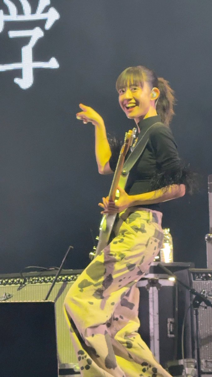 A female guitarist in camouflage pants and a black top performs on stage with an electric guitar, smiling and pointing toward the audience.