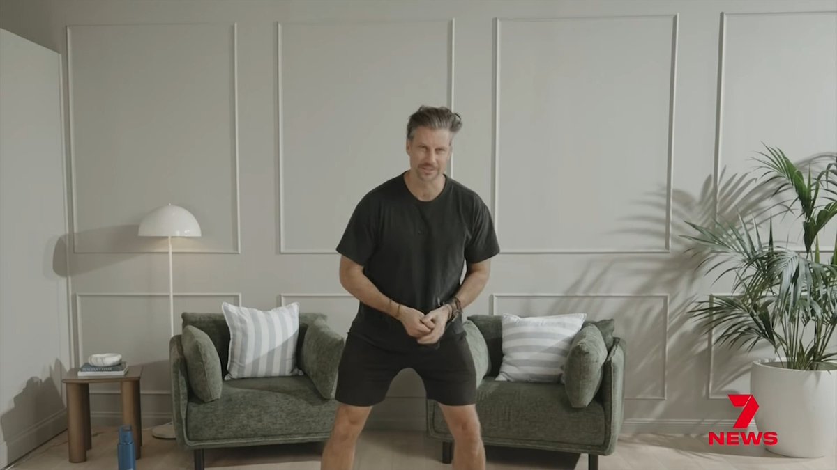 A man in a black t-shirt and shorts stands in a modern living room with a green sofa, striped cushions, a mushroom lamp, and a potted plant, with a "7 NEWS" logo in the corner. The image likely shows Sam Wood, the fitness influencer mentioned in the post who is facing court over domestic violence charges while investors in his former $70 million business absorb financial losses.