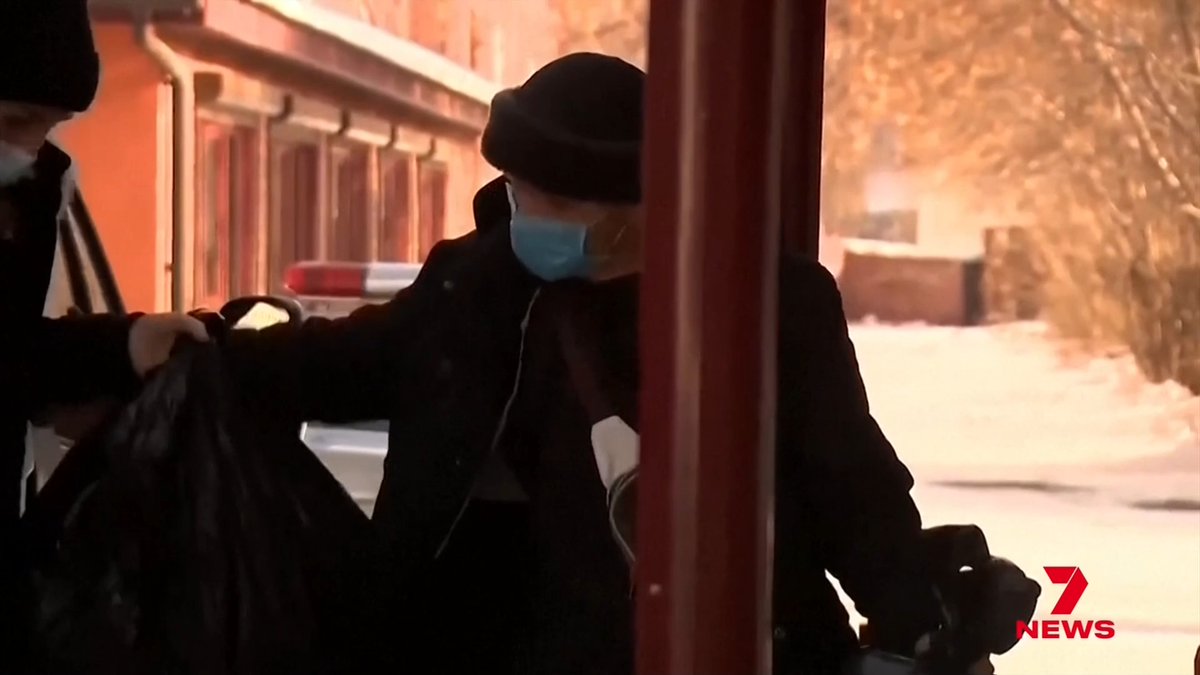 A nighttime news footage from Channel 7 News showing a person wearing a dark winter coat, fur hat, and face mask handling trash bags outside in a snowy environment near a police vehicle.