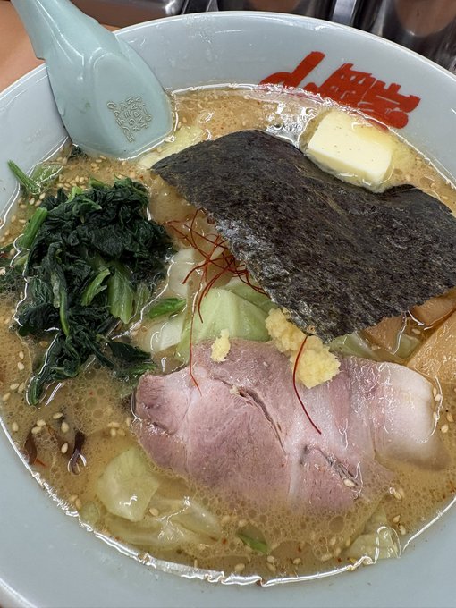 A close-up photograph of a Japanese ramen bowl from J. Men (J麺) featuring pork slices, nori seaweed, spinach, ginger, and butter in a rich broth, with a light blue ceramic spoon.