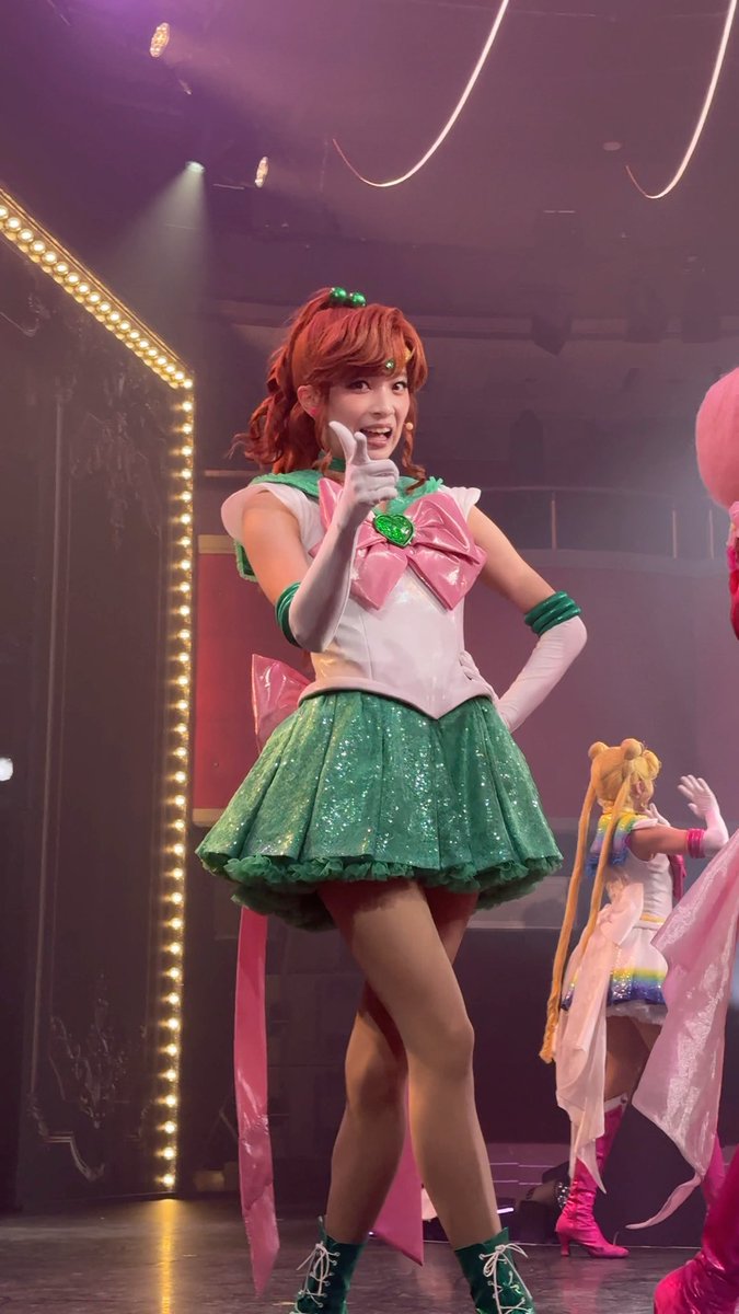 A performer dressed in a green and white Sailor Jupiter costume from Sailor Moon on stage, posing energetically with another Sailor Moon character visible in the background.