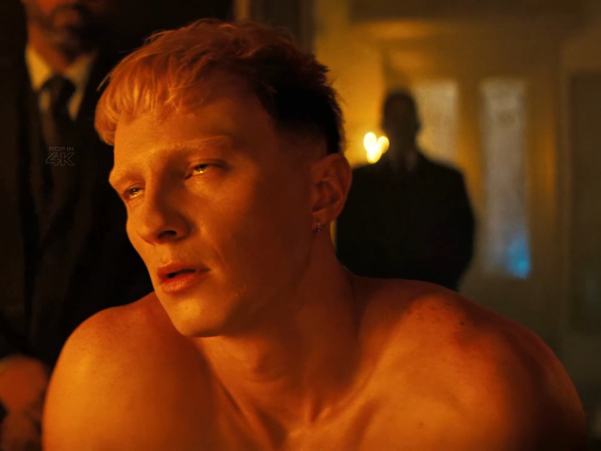 A shirtless person with short reddish hair looks off to the side with a serious expression in a warmly lit room. Text "POP IN 4K" appears on the left side of the frame.