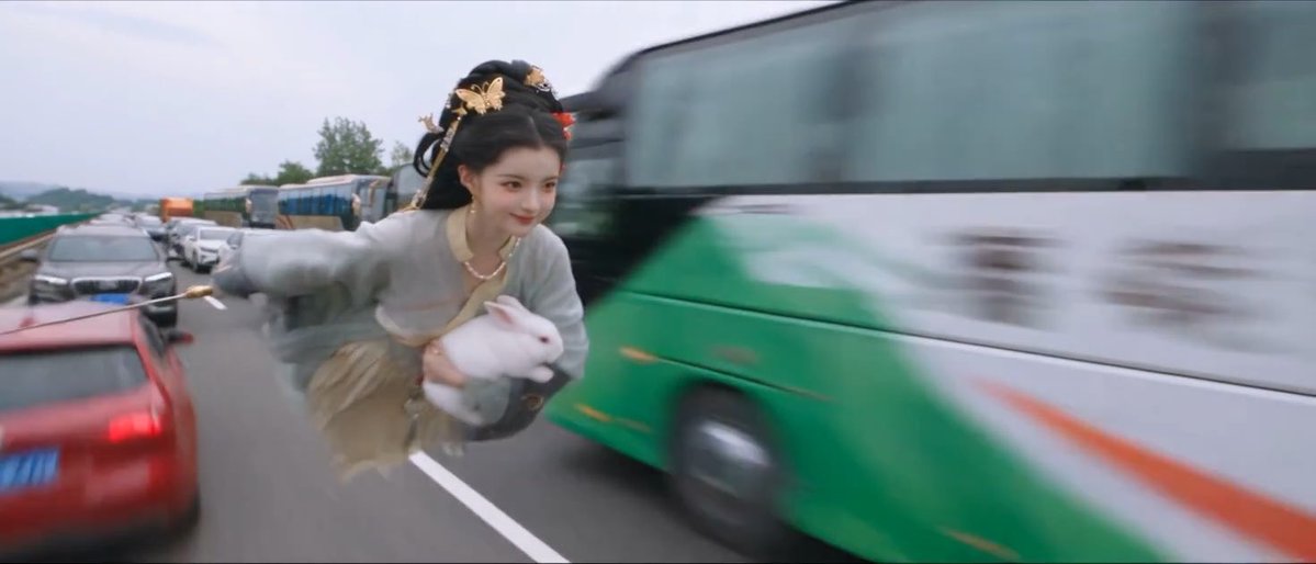 A person in traditional Chinese historical costume holding a white rabbit, digitally edited to appear flying alongside vehicles on a highway next to a green and white bus.