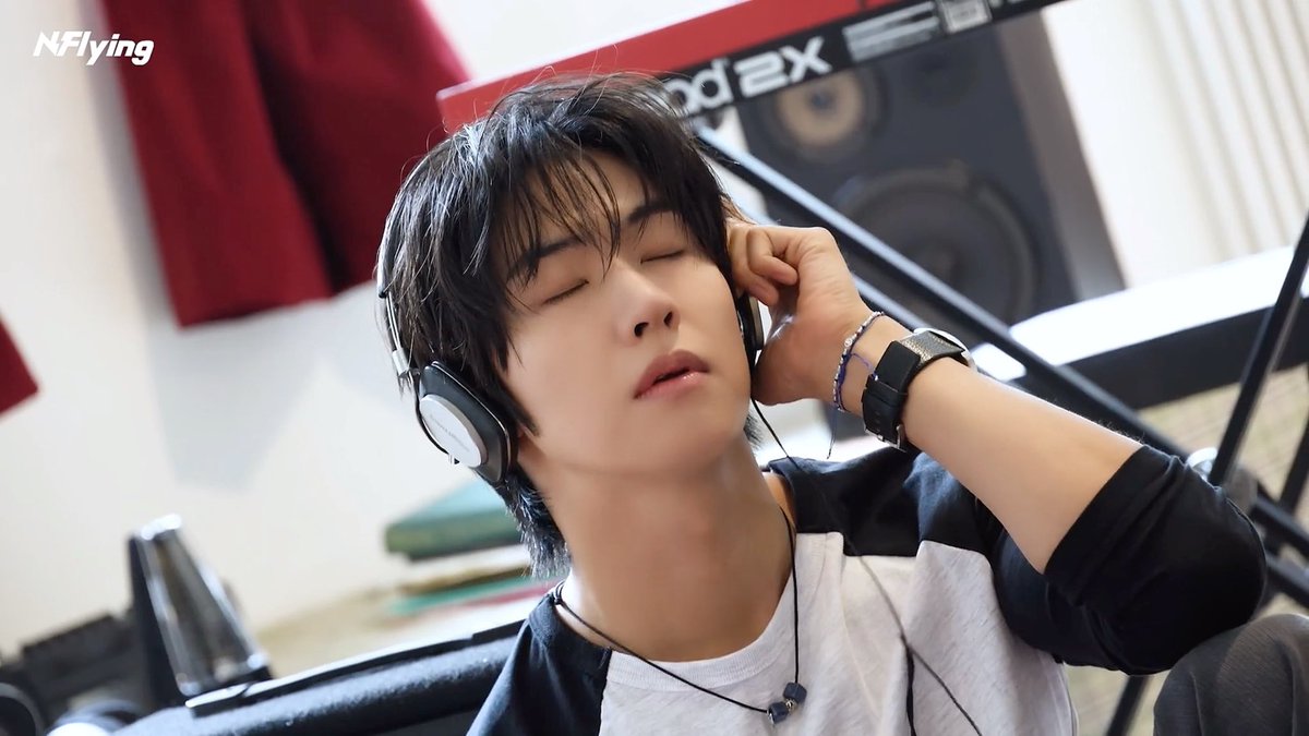 A young man with dark hair listens to music with his eyes closed using headphones in a studio setting, with the 'N.Flying' logo visible in the corner.