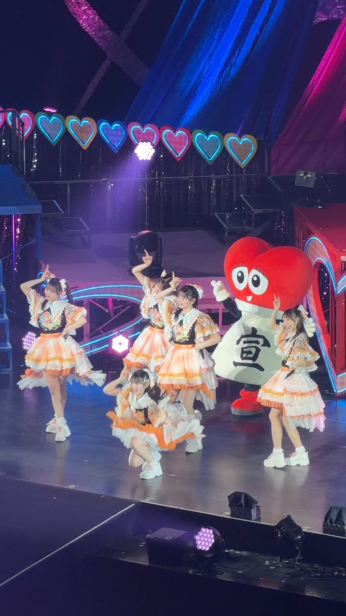 A group of performers in orange and white frilly dresses on stage posing with a large red heart mascot that reads '宮' (miyako).