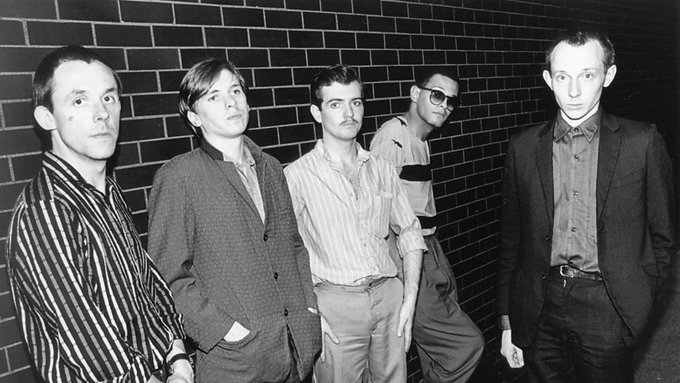 A black-and-white photograph showing five people standing against a brick wall, appearing to be a vintage band or group portrait.