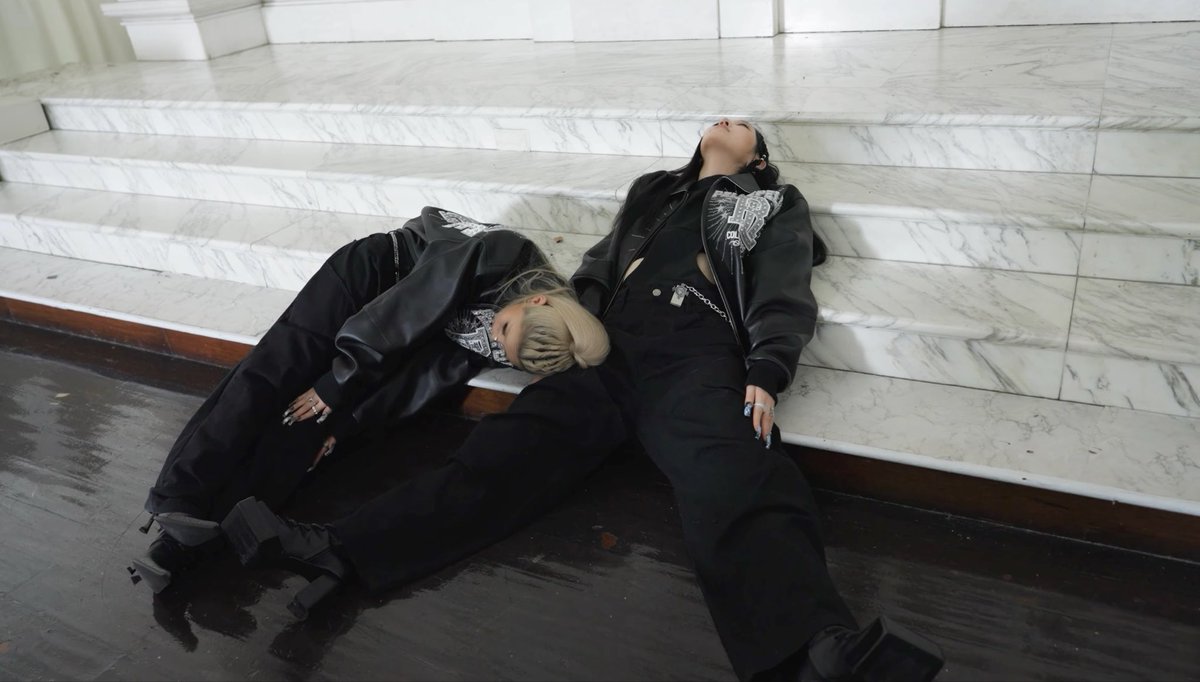 Two people in black streetwear pose dramatically on a white marble staircase, with one figure resting their head on the other's lap.
