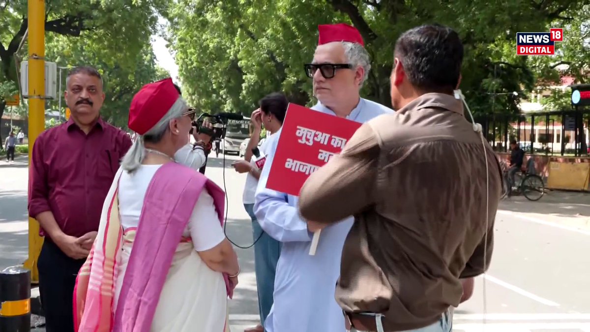 A group of people, including a man in a red cap holding a sign with Hindi text, are gathered outdoors in front of a news camera, with a 'NEWS 18 DIGITAL' logo in the corner.