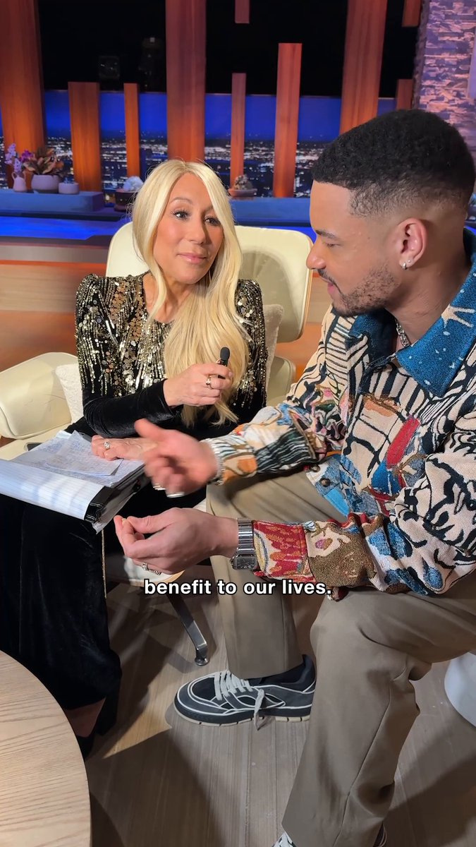 A screenshot from an interview or talk show featuring a blonde woman in a sparkly top speaking with a man in a colorful patterned shirt, with the subtitle 'benefit to our lives.' visible.
