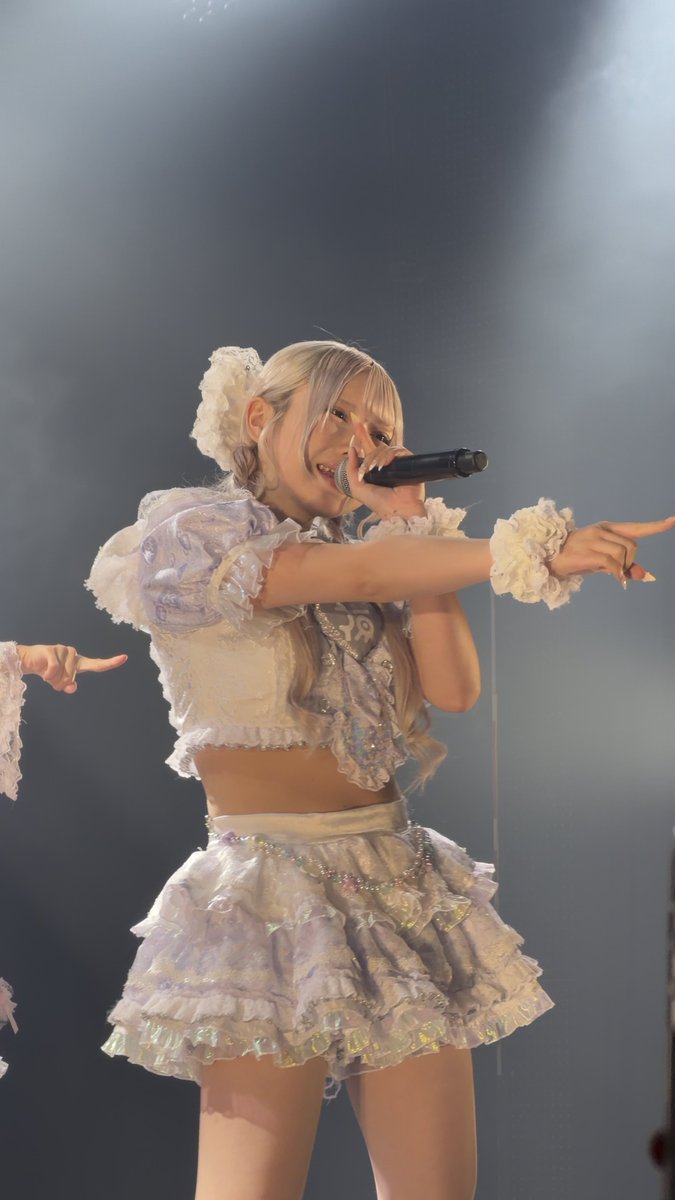A performer with silver-blonde hair in a frilly white and lavender outfit sings into a microphone on stage while pointing, wearing a decorative hair accessory and white wrist cuffs.