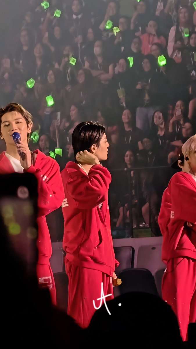 Three performers in matching red outfits stand on a stage with a crowd holding green light sticks behind them; one holds a blue microphone while another adjusts his earpiece.