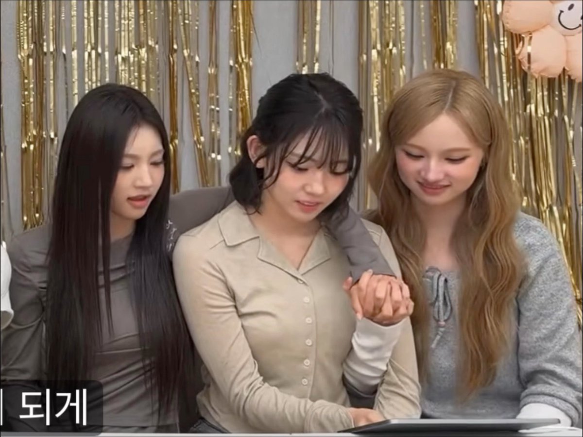 Three young women with long hair, one in grey, one in beige, and one in a grey sweater, posing together and looking down, with the Korean text '되게' visible in the corner.